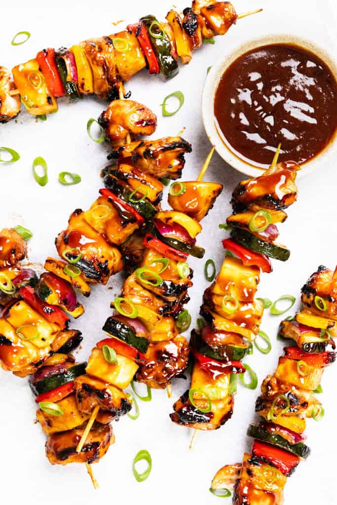 Grilled BBQ chicken and vegetable skewers glazed with sauce, garnished with sliced green onions and pineapple, arranged on a white surface next to a bowl of brown dipping sauce.