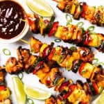 Grilled BBQ chicken and vegetable skewers glazed with sauce, garnished with sliced green onions. Lime wedges, sweet pineapple pieces, and a bowl of dipping sauce are arranged on a white surface.