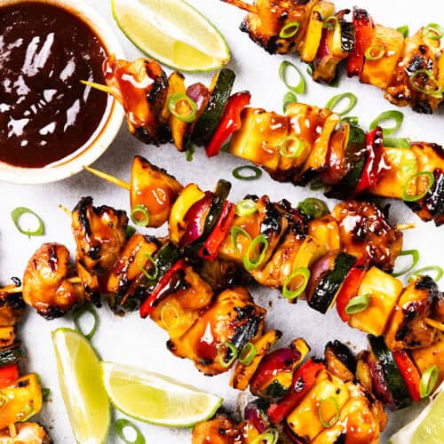 Grilled BBQ chicken and vegetable skewers glazed with sauce, garnished with sliced green onions. Lime wedges, sweet pineapple pieces, and a bowl of dipping sauce are arranged on a white surface.