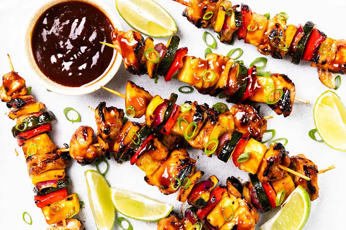 Grilled BBQ chicken and vegetable skewers glazed with sauce, garnished with sliced green onions. Lime wedges, sweet pineapple pieces, and a bowl of dipping sauce are arranged on a white surface.