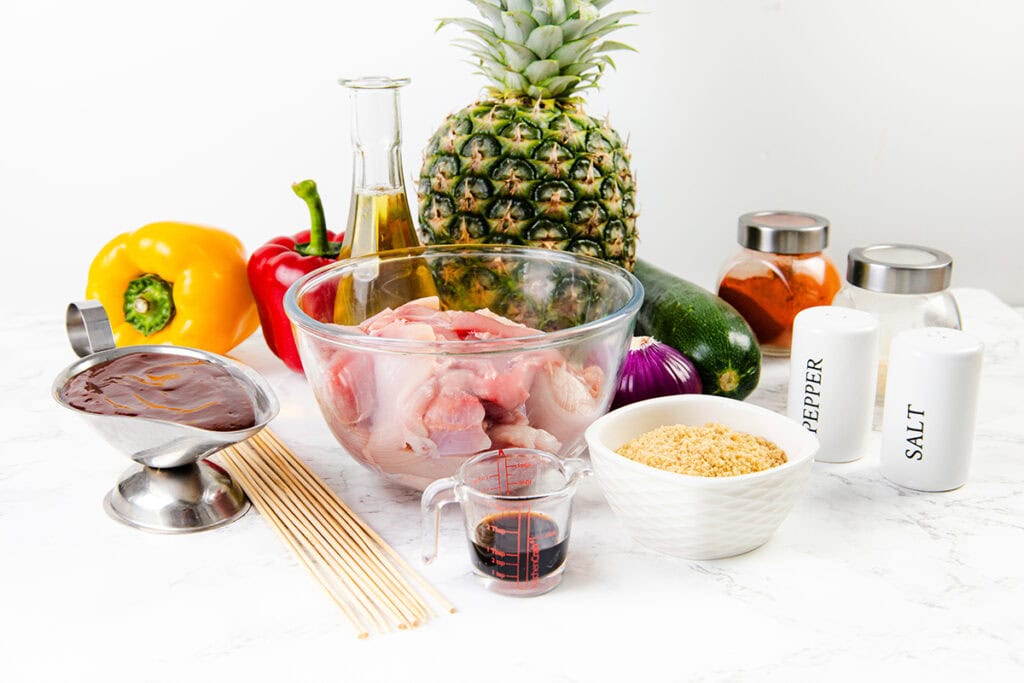 A variety of ingredients for a recipe, including raw chicken in a glass bowl, pineapple, colorful bell peppers, zucchini, red onion, barbecue sauce for BBQ chicken skewers, soy sauce, breadcrumbs, oil, spices, salt, pepper, and wooden skewers.