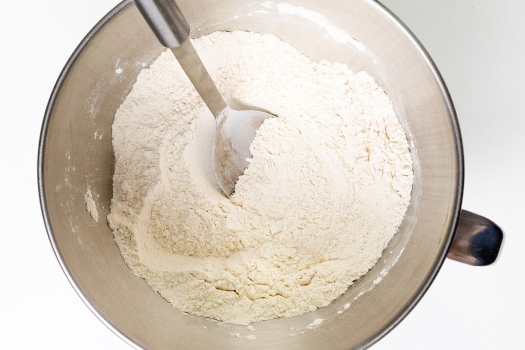A metal mixing bowl filled with flour, ready for a Basic Pizza Dough, with a metal spoon partially buried in the flour, viewed from above on a white background.