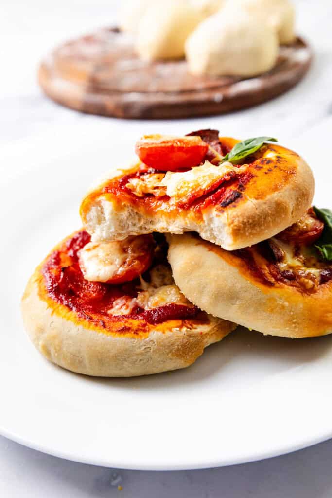 Three mini pizzas made with homemade pizza dough are topped with tomato sauce, melted cheese, cherry tomatoes, and fresh basil, stacked on a white plate. In the background, a wooden board displays blocks of cheese.