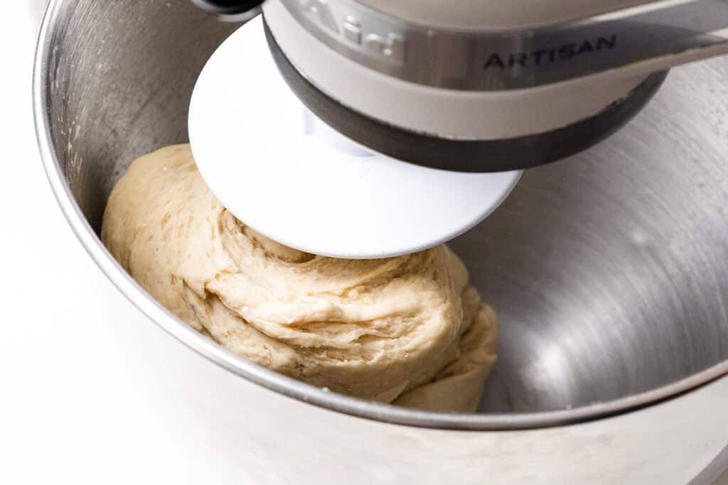 Close-up of basic pizza dough being kneaded in a stainless steel stand mixer bowl using a white dough hook attachment.