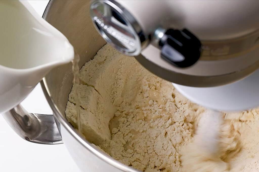 A close-up of a stand mixer blending flour in a metal bowl while liquid is poured in from a white pitcher—perfect for making homemade pizza dough.