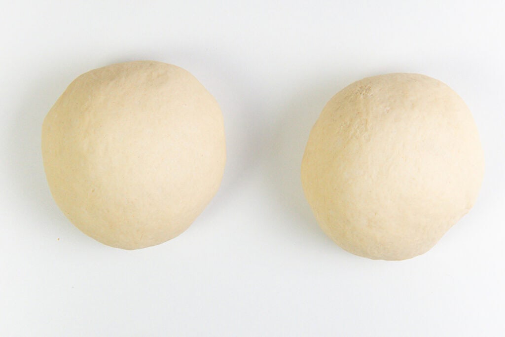 Two round balls of homemade pizza dough are placed side by side on a white surface.