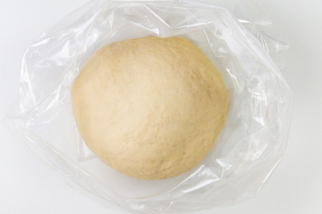 A round ball of homemade pizza dough sits in the center of a clear plastic bag, resting on a white surface.