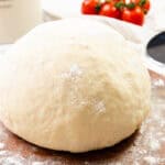 A ball of pizza dough dusted with flour sits on a wooden surface, ready for your homemade pizza masterpiece, with tomatoes and a pizza cutter in the background.