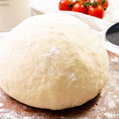 A ball of pizza dough dusted with flour sits on a wooden surface, ready for your homemade pizza masterpiece, with tomatoes and a pizza cutter in the background.