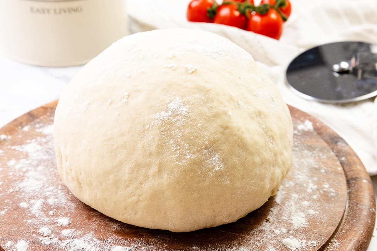 A ball of pizza dough dusted with flour sits on a wooden surface, ready for your homemade pizza masterpiece, with tomatoes and a pizza cutter in the background.