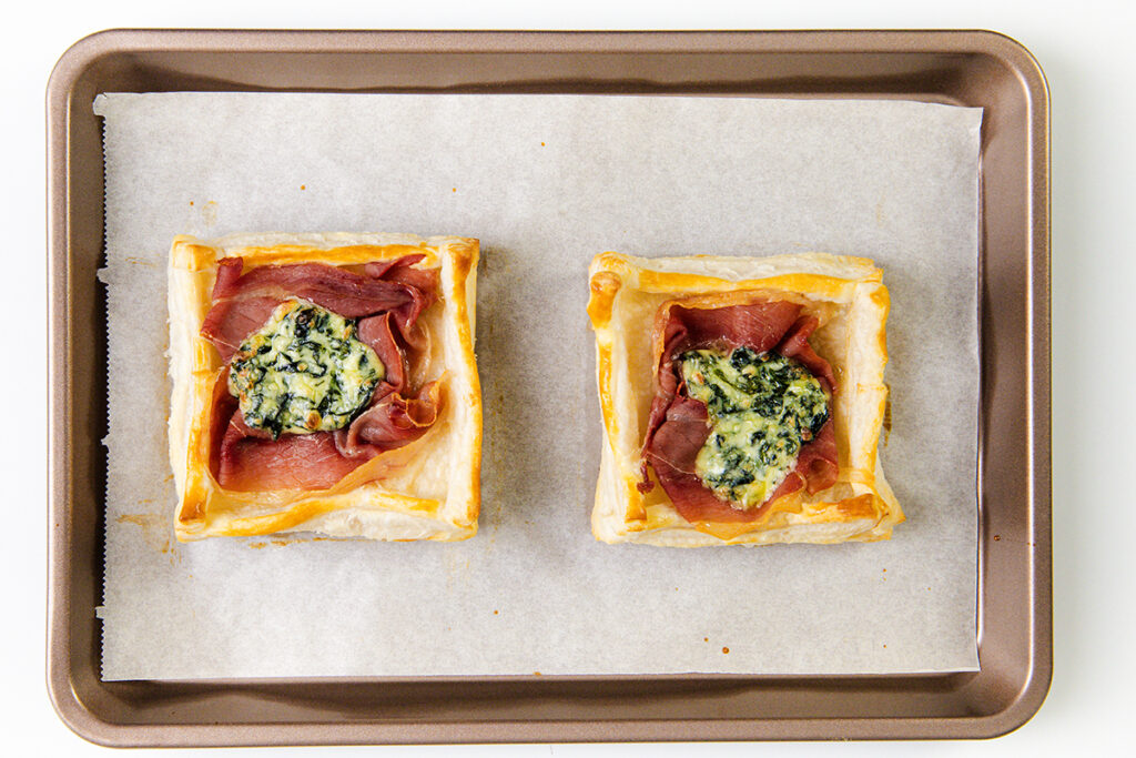 Two Puff Pastry breakfast squares filled with prosciutto and a dollop of spinach and cheese mixture, baked to perfection on a parchment-lined baking sheet.