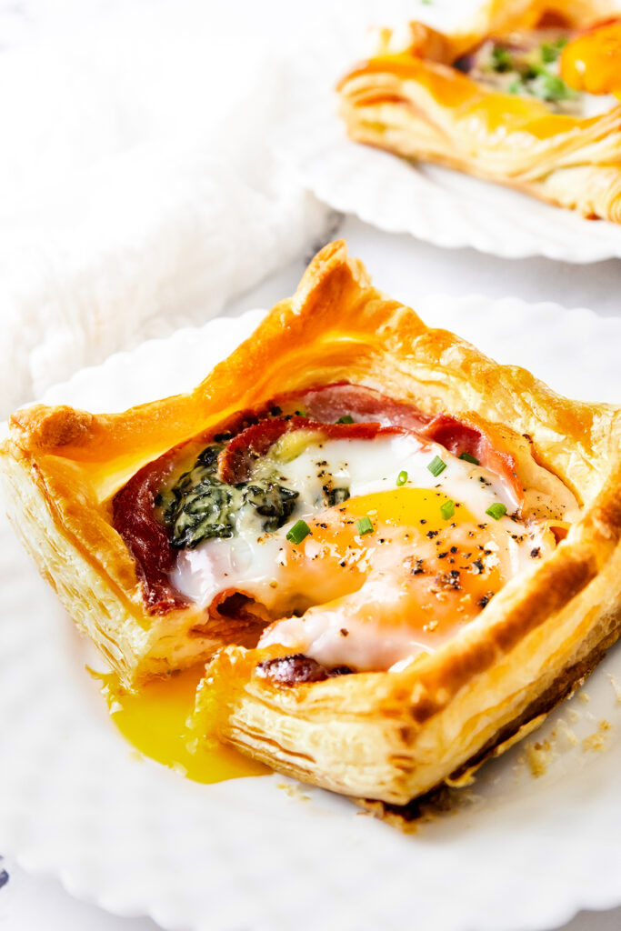 A square Puff Pastry Breakfast tart filled with egg, spinach, and prosciutto, garnished with chives and black pepper. The runny yolk spills onto a white plate, with another tempting tart in the background.
