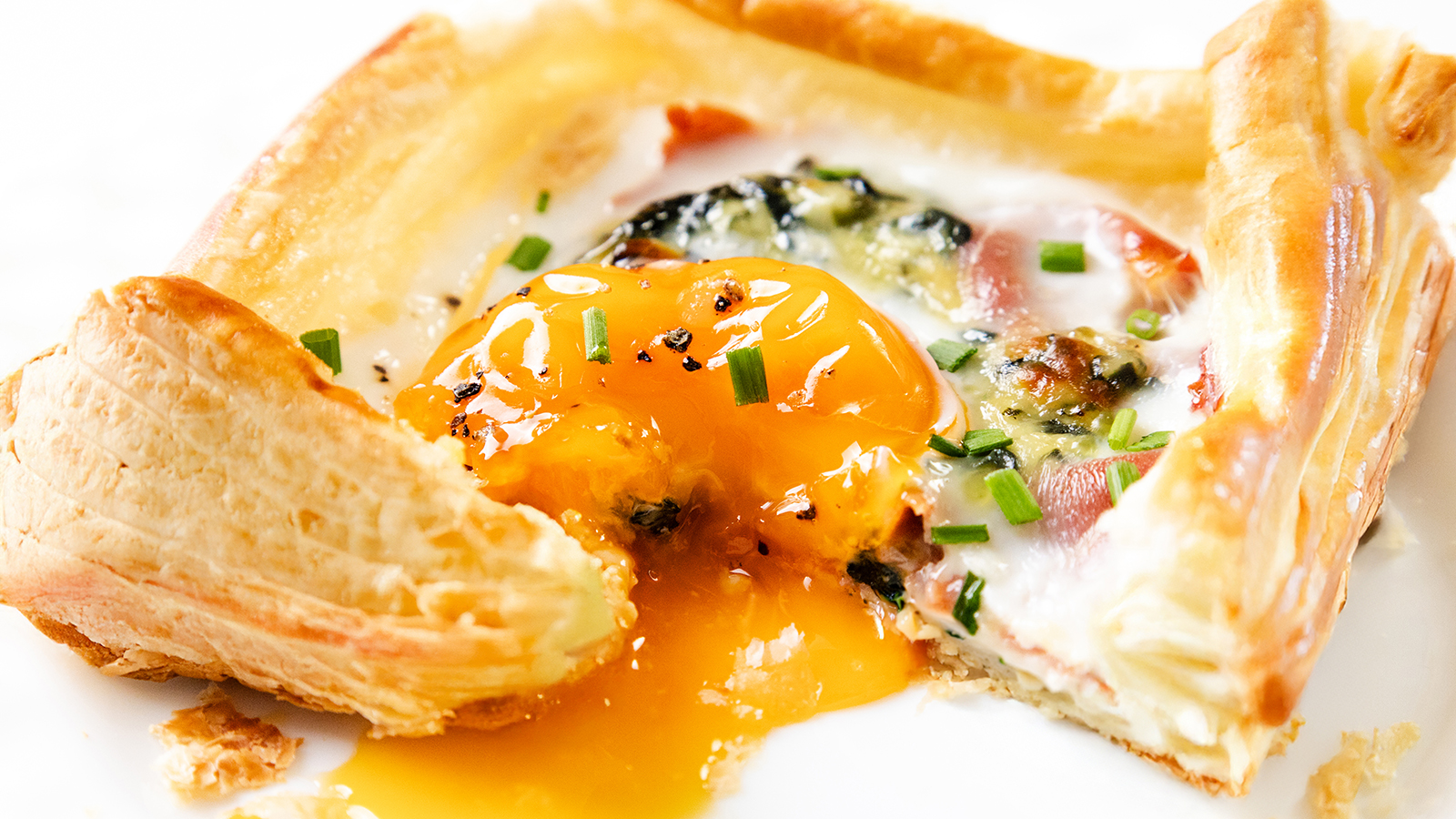 A flaky pastry square filled with a sunny-side-up egg, chopped herbs, and cheese; the yolk is broken and oozing onto the plate—perfect for a special Mother’s Day Brunch.