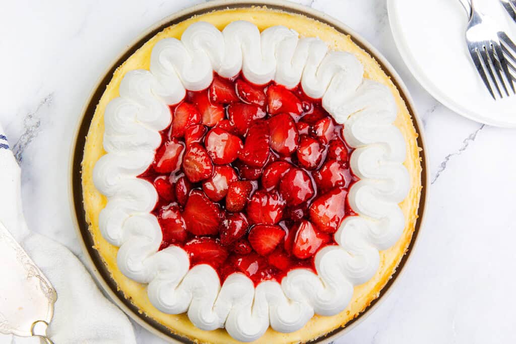 A creamy strawberry cheesecake topped with glazed strawberries in the center and a thick ring of piped whipped cream around the edge, placed on a marble surface next to plates and forks.