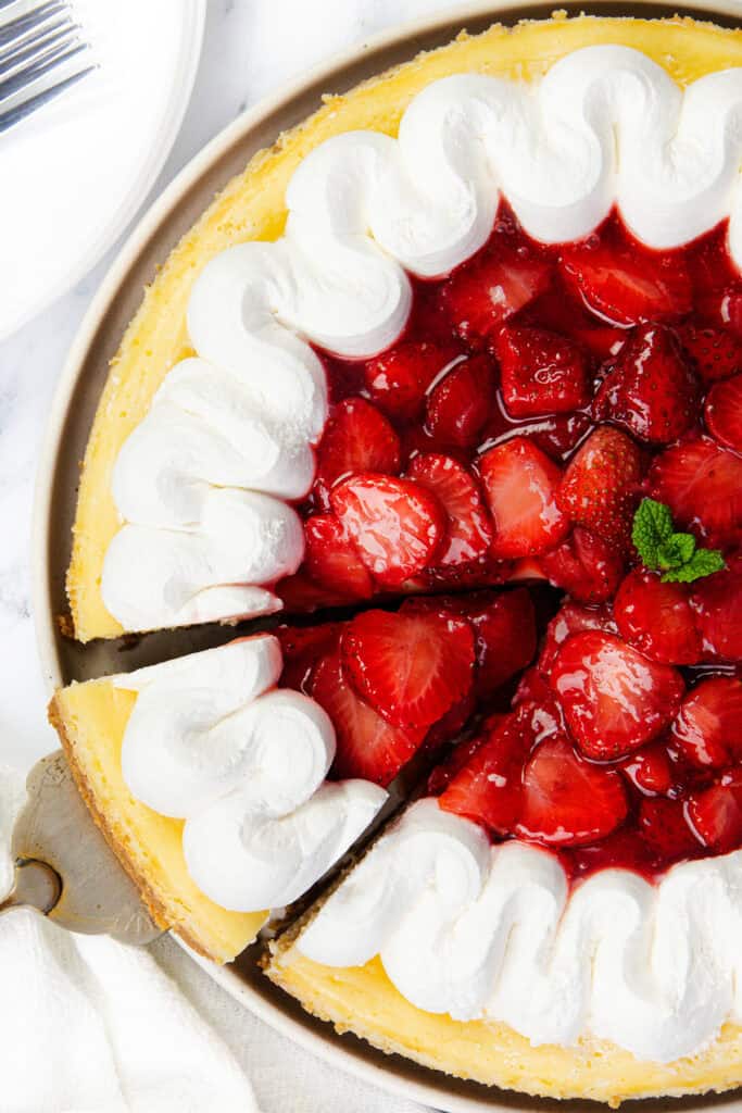 A top view of a creamy strawberry cheesecake topped with glazed strawberries and dollops of whipped cream around the edge, with one slice partially removed. A sprig of mint decorates the center.