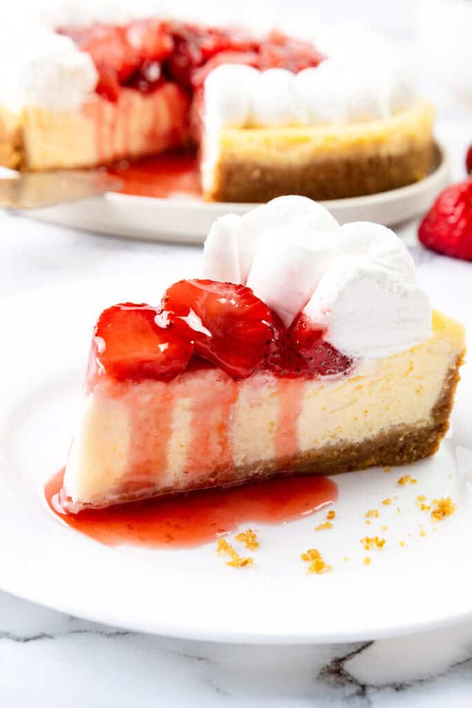 A slice of creamy strawberry cheesecake with a graham cracker crust, topped with strawberry sauce and whipped cream, sits on a white plate. The whole cheesecake is in the background with fresh strawberries nearby.