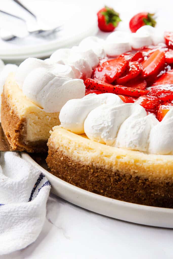A creamy cheesecake with a graham cracker crust, topped with whipped cream and fresh sliced strawberries, sits on a white plate. A slice of this luscious strawberry cheesecake has been cut, with extra strawberries and plates in the background.