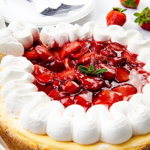 A creamy cheesecake topped with glazed strawberries and a ring of whipped cream, garnished with a mint sprig. Fresh strawberries and stacked plates with forks add the perfect touch in the background.