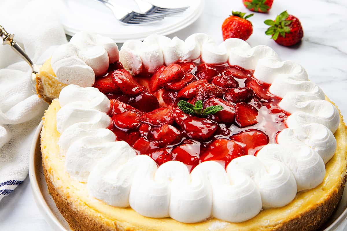 A creamy cheesecake topped with glazed strawberries and a ring of whipped cream, garnished with a mint sprig. Fresh strawberries and stacked plates with forks add the perfect touch in the background.