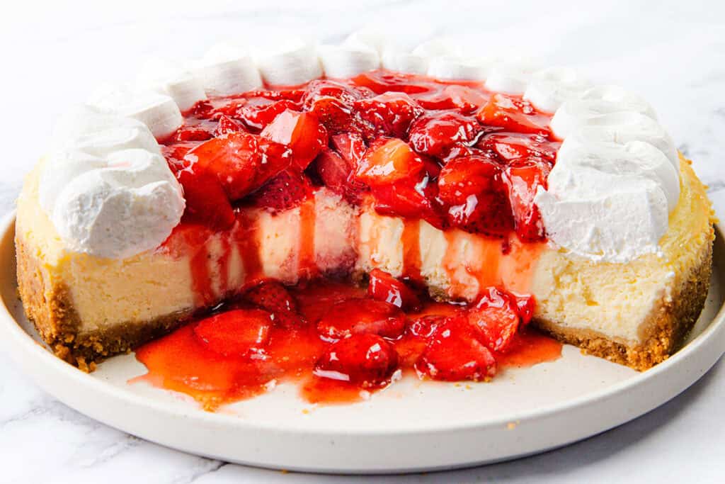 A creamy cheesecake topped with strawberry sauce and whole strawberries, decorated with whipped cream rosettes around the edge, is displayed on a white plate with a slice missing to reveal its luscious interior.