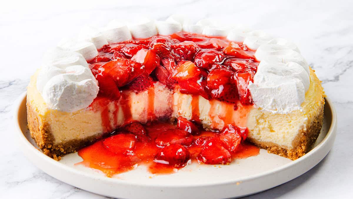 A classic among strawberry desserts, this cheesecake is topped with glazed strawberries and surrounded by dollops of whipped cream on a white plate, with a large slice removed to reveal its creamy interior and crumbly crust.