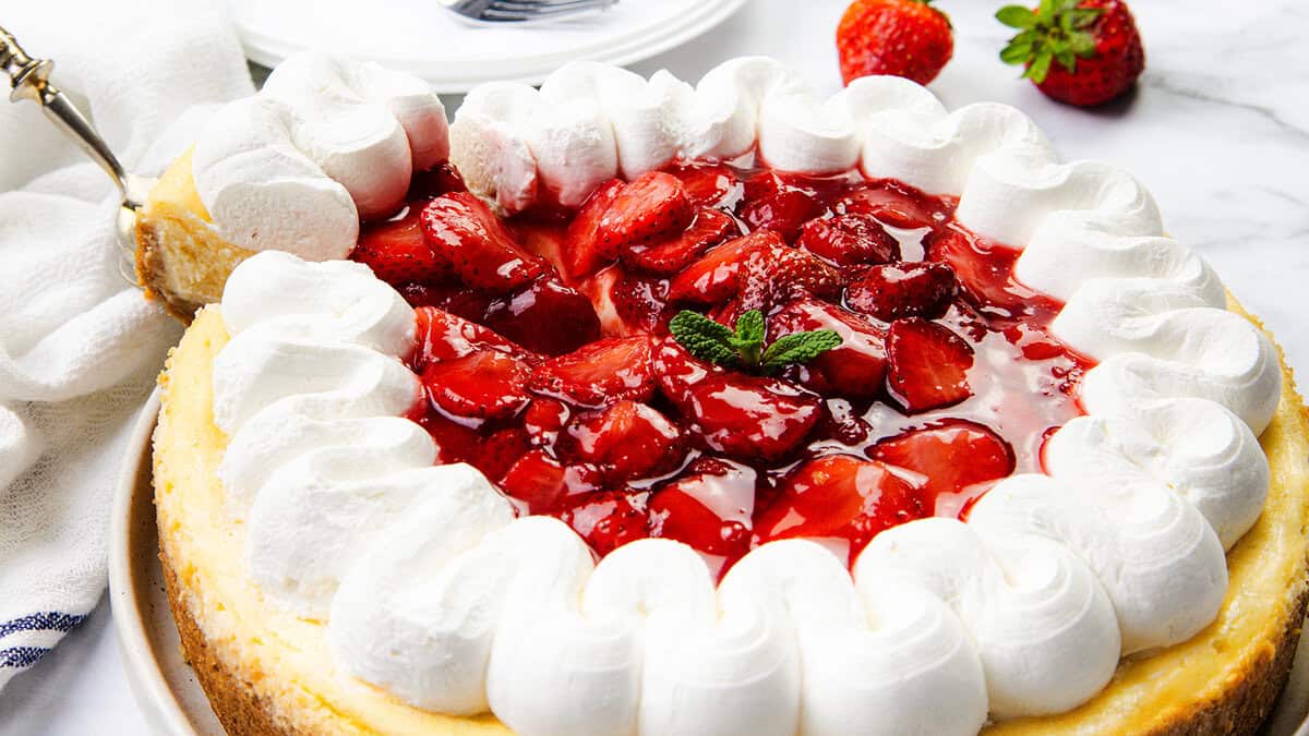 A cheesecake topped with glossy strawberry sauce and fresh strawberries, surrounded by piped swirls of whipped cream, sits on a white surface—perfect inspiration for your next Dessert Recipes adventure.