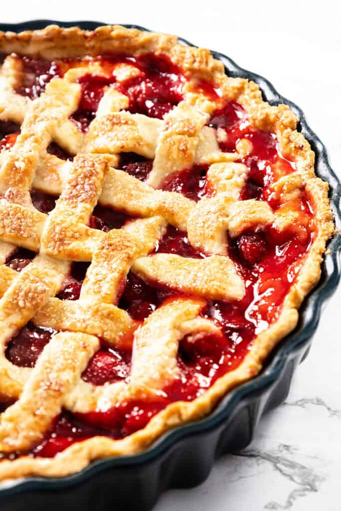 A close-up of a freshly baked cherry pie with a golden, flaky butter crust and glossy red filling, in a dark pie dish on a white surface.