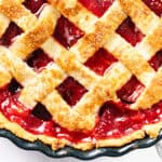 A close-up of a cherry pie with a flaky butter crust and glossy red filling, in a round pie dish on a white surface.