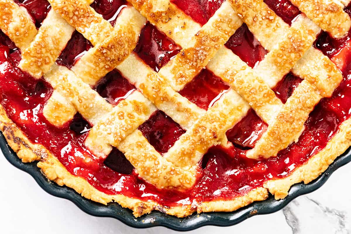 A close-up of a cherry pie with a flaky butter crust and glossy red filling, in a round pie dish on a white surface.