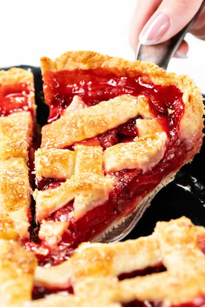 A close-up of a hand lifting a slice of cherry pie with a flaky butter crust from a pie dish. The bright red, glossy filling contrasts beautifully with the golden, sugar-sprinkled crust.