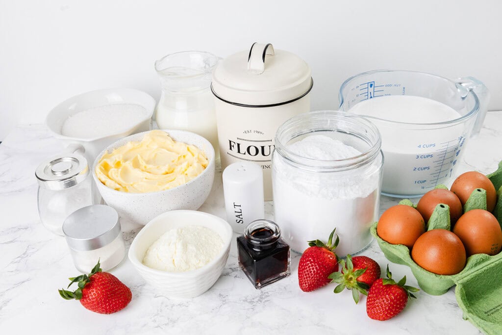 Various baking ingredients for Strawberry Shortcake, including flour, sugar, butter, eggs, vanilla extract, milk, salt, baking powder, powdered sugar, and fresh strawberries arranged on a white countertop.