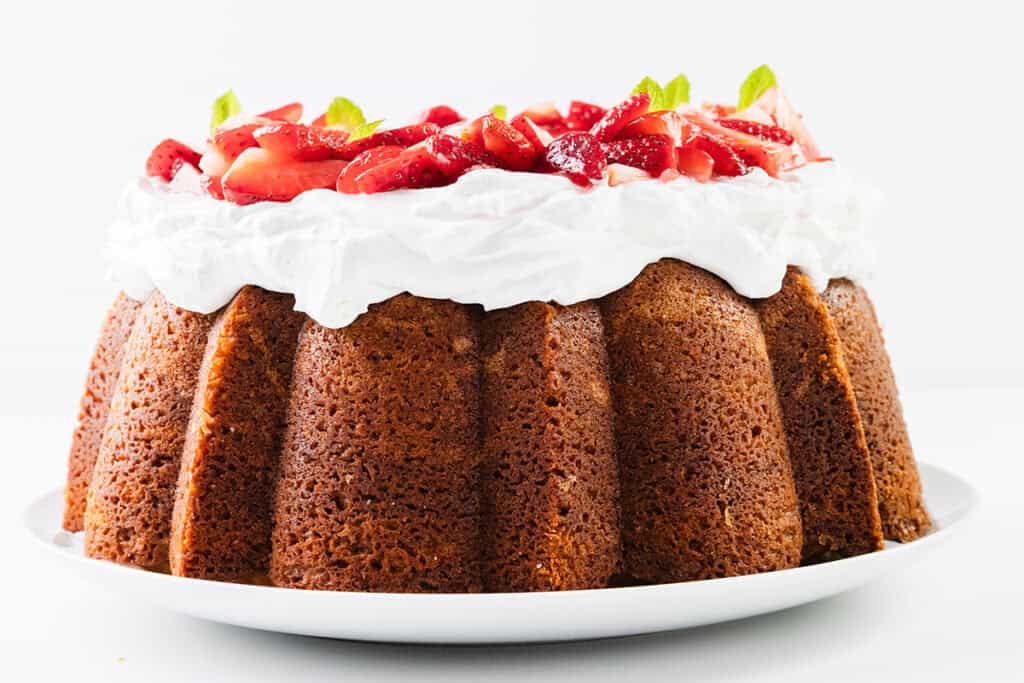 A golden brown Strawberry Shortcake Bundt Cake topped with a thick layer of whipped cream, fresh sliced strawberries, and mint leaves, displayed on a white plate against a white background.