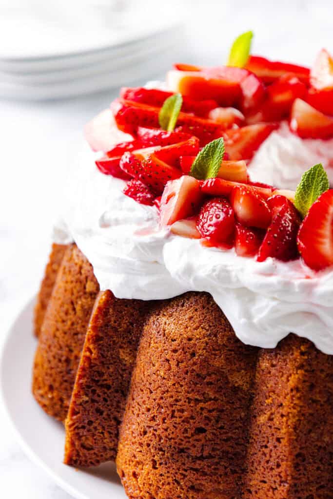 A Strawberry Bundt Cake topped with a generous layer of whipped cream, fresh sliced strawberries, and small mint leaves is displayed on a white plate. The background is softly blurred with stacked plates visible.