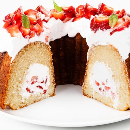 A Strawberry Shortcake Bundt Cake with a cream and strawberry filling is topped with whipped cream, sliced strawberries, and mint leaves, sitting on a white plate. A large slice has been cut to reveal the delicious filled bundt cake center.