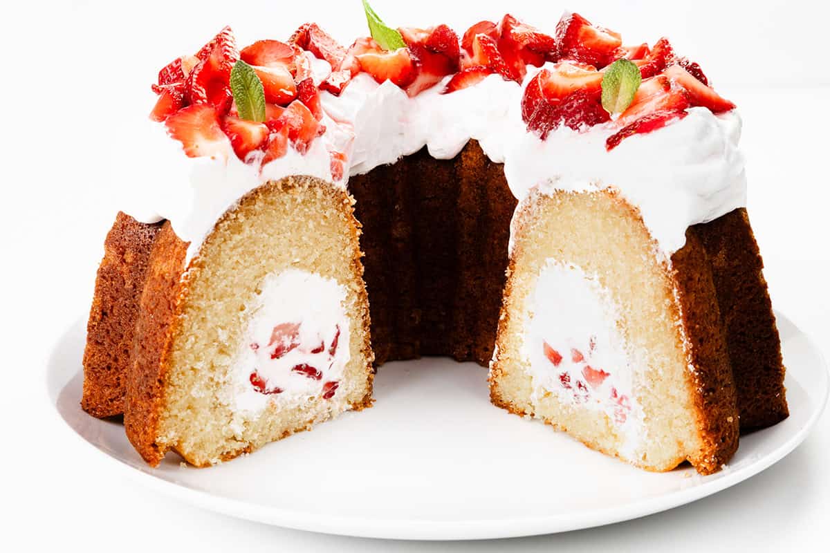 A Strawberry Shortcake Bundt Cake with a cream and strawberry filling is topped with whipped cream, sliced strawberries, and mint leaves, sitting on a white plate. A large slice has been cut to reveal the delicious filled bundt cake center.