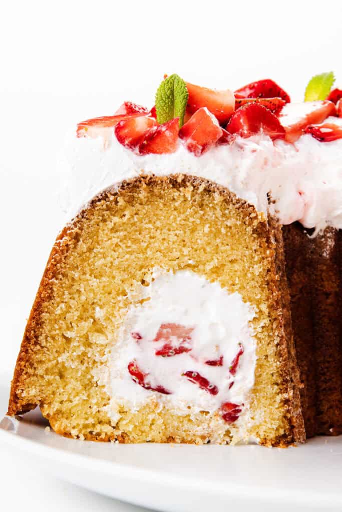 A close-up of a sliced Bundt Cake inspired by strawberry shortcake, filled with whipped cream and strawberries, then topped with more whipped cream, fresh strawberry pieces, and mint leaves.