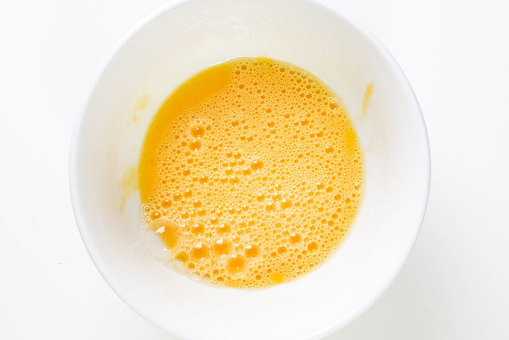 A white bowl filled with beaten eggs, showing a frothy, yellow mixture with bubbles on the surface—perfect for creating a delicious filled cake—set against a plain white background.