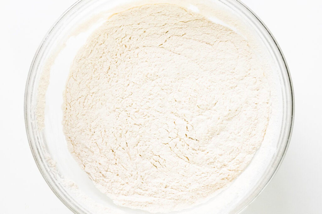 A clear glass bowl filled with a mound of dry, white flour, seen from above on a white background—perfect for baking Bundt Cake or Strawberry Shortcake.