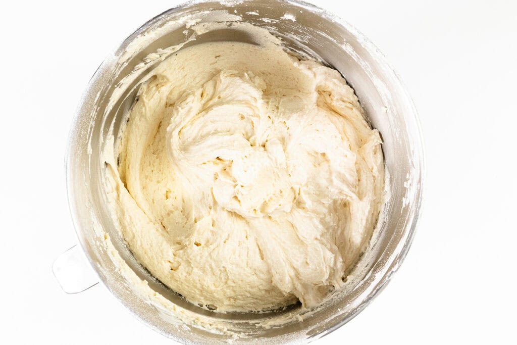 A metal mixing bowl filled with creamy, light-colored strawberry shortcake batter, viewed from above against a white background.