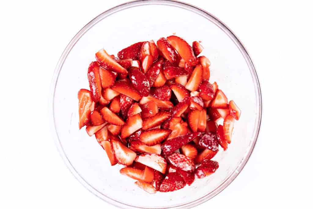 A glass bowl filled with sliced strawberries coated in sugar, reminiscent of a classic strawberry shortcake, set against a white background.