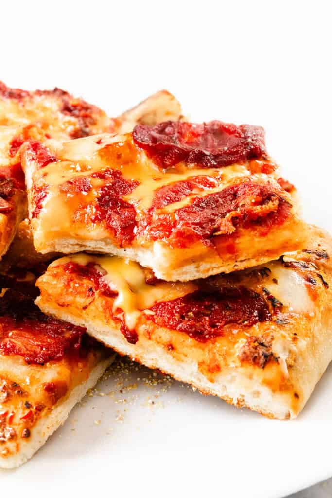 Close-up of thick, rectangular slices of Trenton Tomato Pie with melted cheese and tomato sauce, stacked on a white plate. The airy, golden brown crust highlights this classic style.