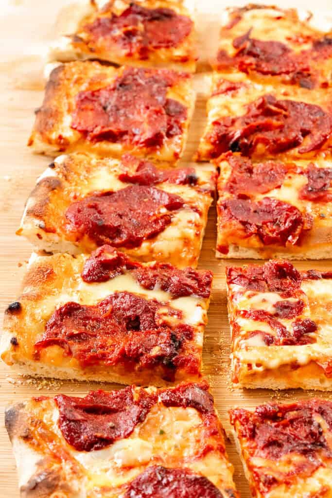 Nine rectangular slices of Trenton Tomato Pie with a crispy, slightly charred crust, melted cheese, and pepperoni on a wooden surface.