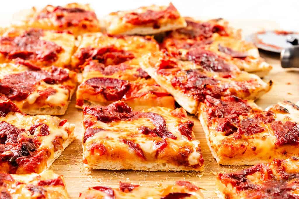 Rectangular slices of thick, homemade Trenton Tomato Pie with melted cheese and rich tomato sauce are arranged on a wooden cutting board. The golden crust shines and some pizza slices are slightly overlapping for a classic, rustic touch.
