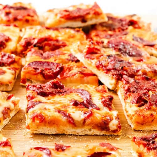 Rectangular slices of thick, homemade Trenton Tomato Pie with melted cheese and rich tomato sauce are arranged on a wooden cutting board. The golden crust shines and some pizza slices are slightly overlapping for a classic, rustic touch.