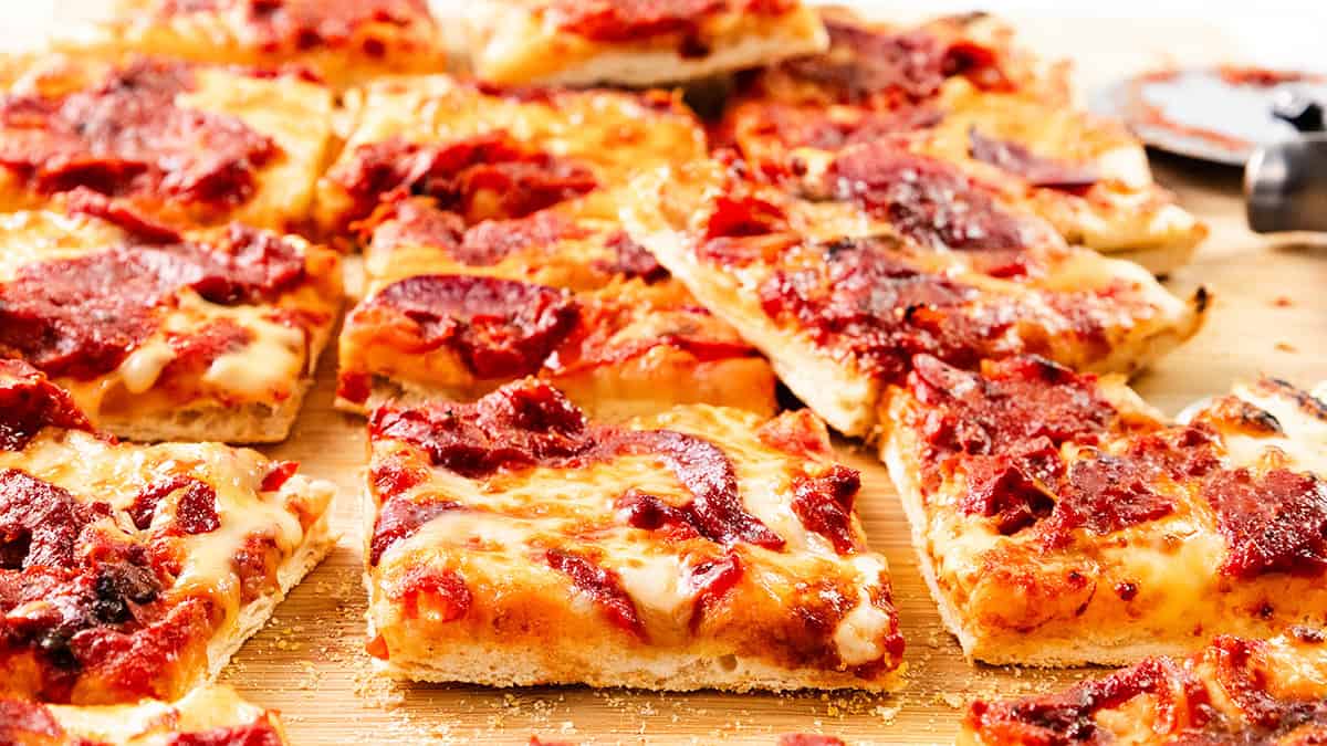 Several square slices of Sicilian-style pizza with melted cheese, tomato sauce, and pepperoni are arranged on a wooden surface—a delicious addition to your collection of comfort food recipes. A pizza cutter is partially visible in the background.