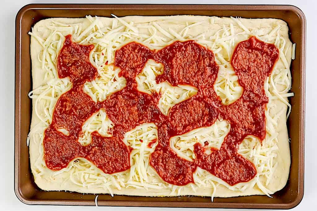 A rectangular sheet pan pizza reminiscent of a classic Trenton Tomato Pie, featuring an uncooked crust, shredded cheese, and unevenly spread tomato sauce—this pizza is ready to be baked.