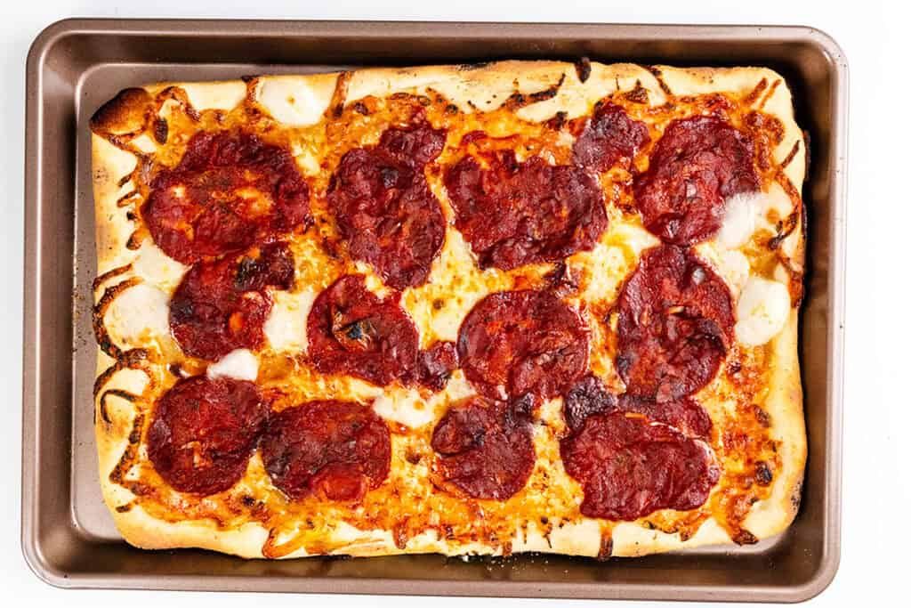 A rectangular pizza with melted cheese and a golden crust sits in a metal baking pan, viewed from above. Reminiscent of a classic Trenton Tomato Pie, pepperoni slices are spread unevenly over the surface.