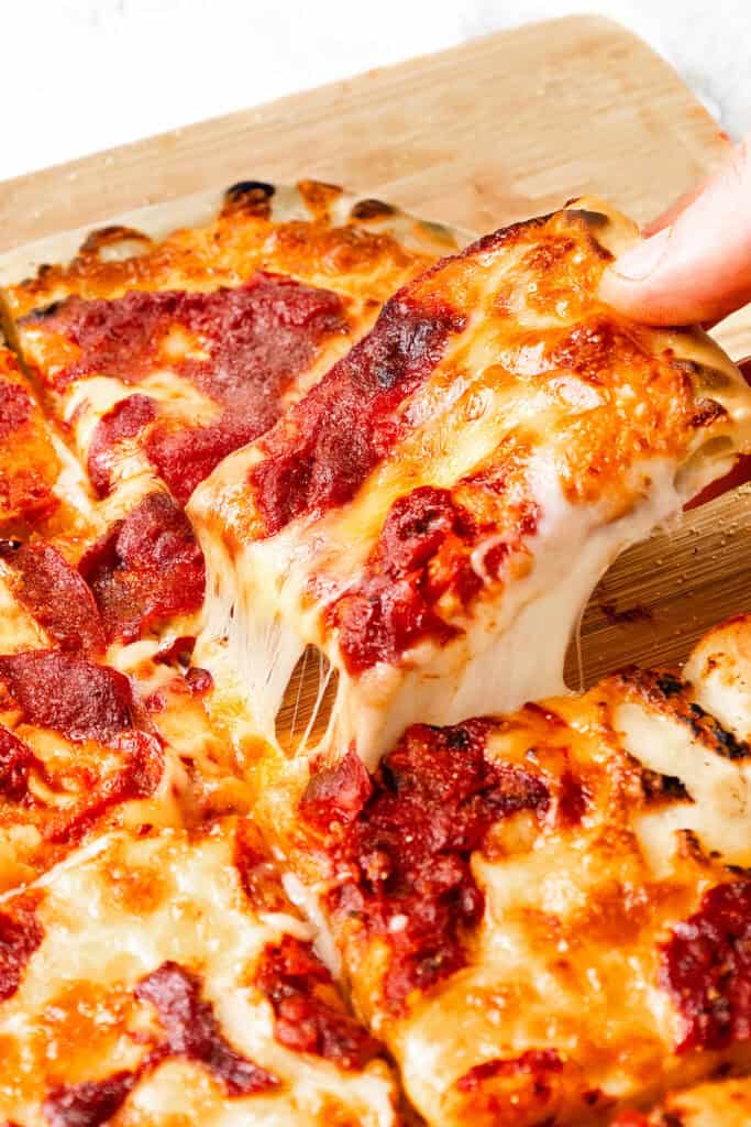A close-up of a hand lifting a cheesy slice of freshly baked Trenton Tomato Pie from a wooden cutting board, with gooey melted cheese stretching from the pizza.