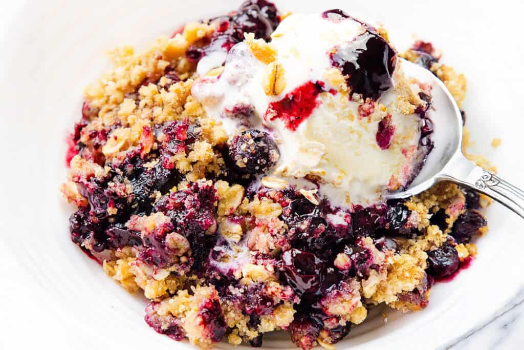 A close-up of a Blueberry Crumble dessert topped with a scoop of melting vanilla ice cream on a white plate, with a spoon partially in the dish.
