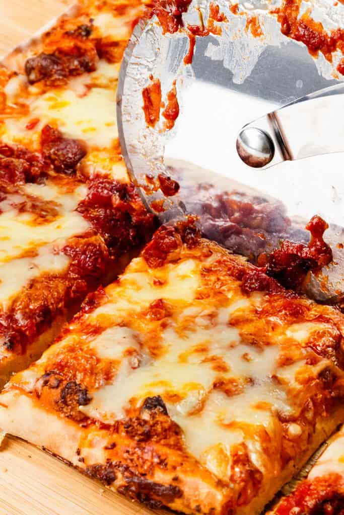 A close-up of a pizza cutter slicing through a cheesy, rectangular homemade pizza with tomato sauce and melted mozzarella on a wooden surface.